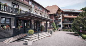 Hoteleingang vom H24 Hotel Hochfirst mit Treppe und Rampe, Topfpflanzen im Freien, Balkone mit Blumenkästen und Nebengebäude mit Holzelementen.