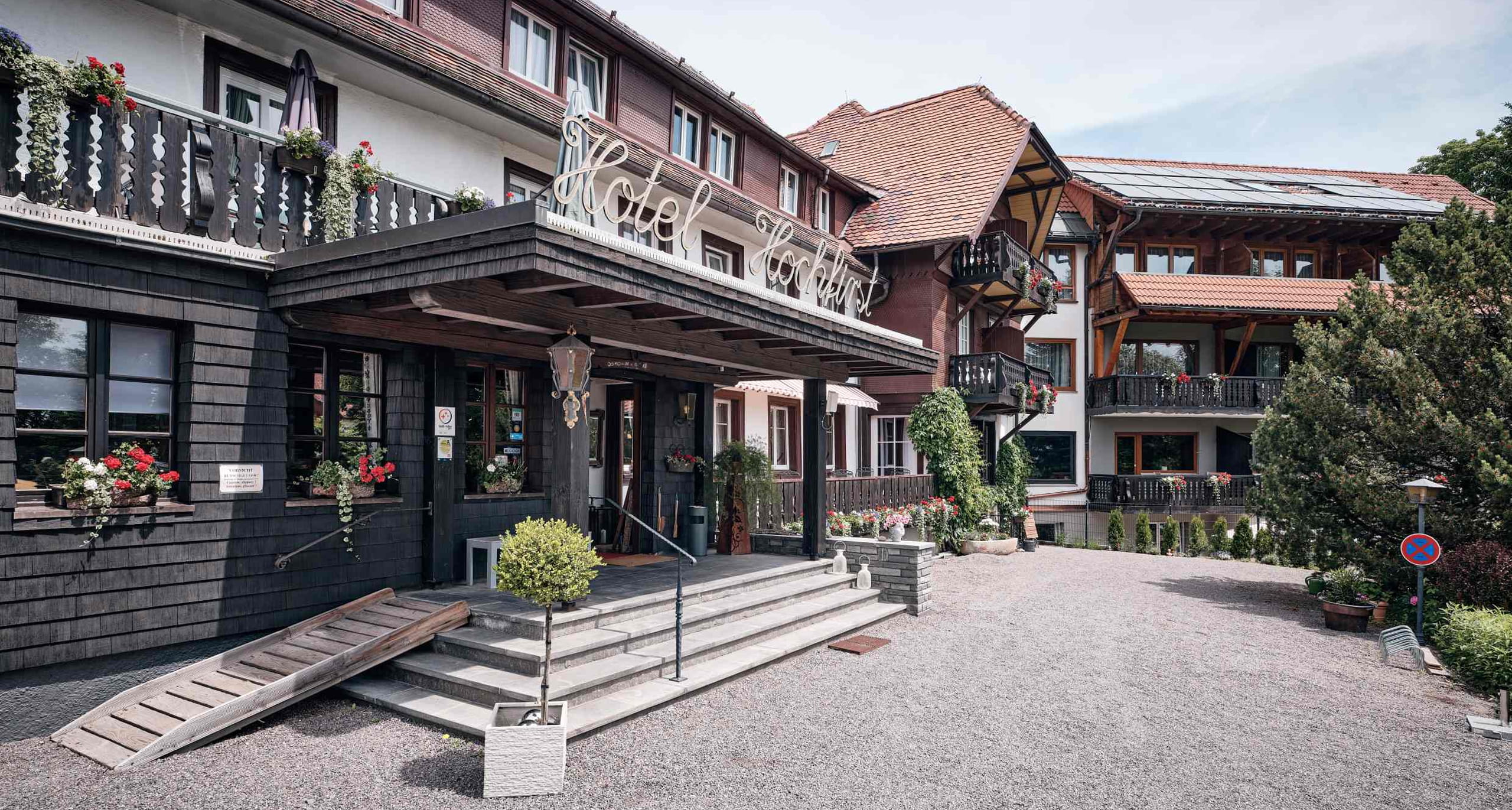 Hoteleingang vom H24 Hotel Hochfirst mit Treppe und Rampe, Topfpflanzen im Freien, Balkone mit Blumenkästen und Nebengebäude mit Holzelementen.