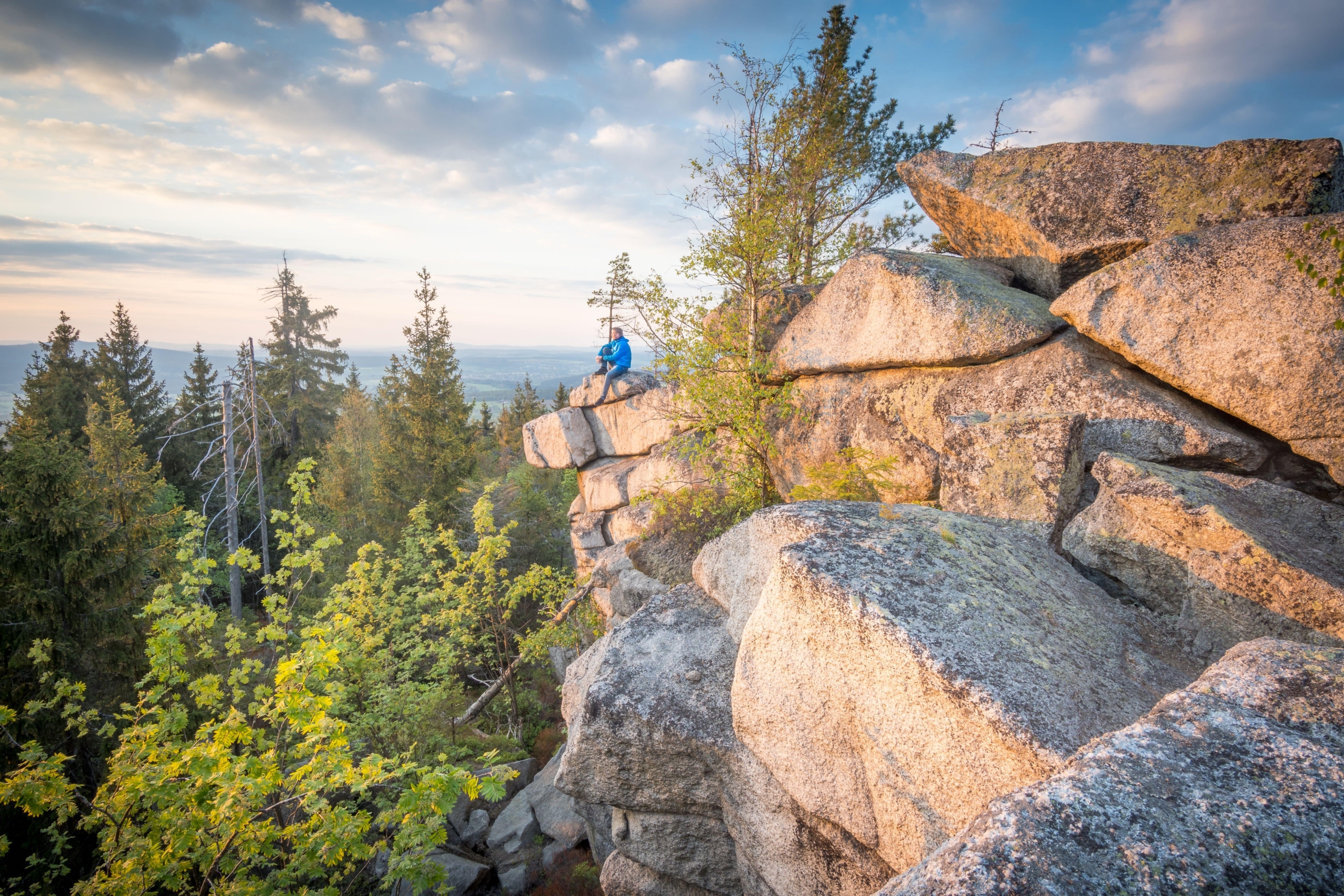 Eine Person in einer blauen Jacke sitzt auf einem von Bäumen umgebenen Felsvorsprung und überblickt eine bewaldete Landschaft im Harz bei Thale unter einem teilweise bewölkten Himmel.