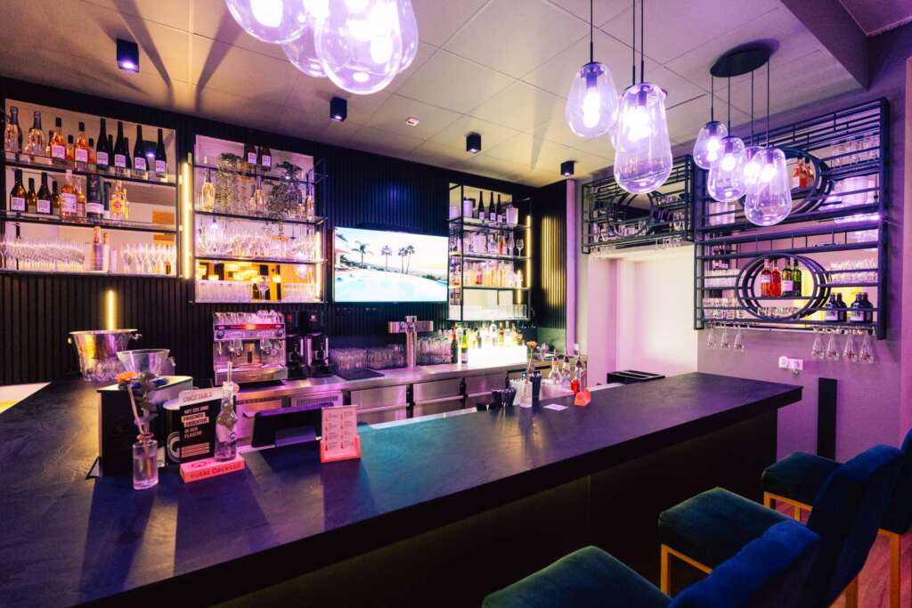 H24 Hotelbar mit schwarzem Tresen, Flaschenregalen und lila Ambiente