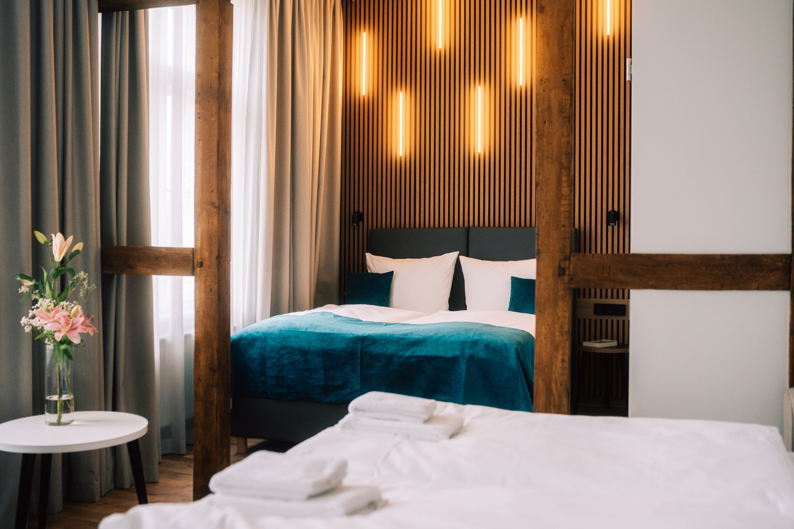 Modernes Hotelzimmer mit einem Doppelbett, tealfarbener Bettwäsche, vertikalen Wandleuchten, Holzlatten, einer Vase mit Blumen auf einem kleinen Tisch und gefalteten Handtüchern auf einem anderen Bett im Vordergrund.