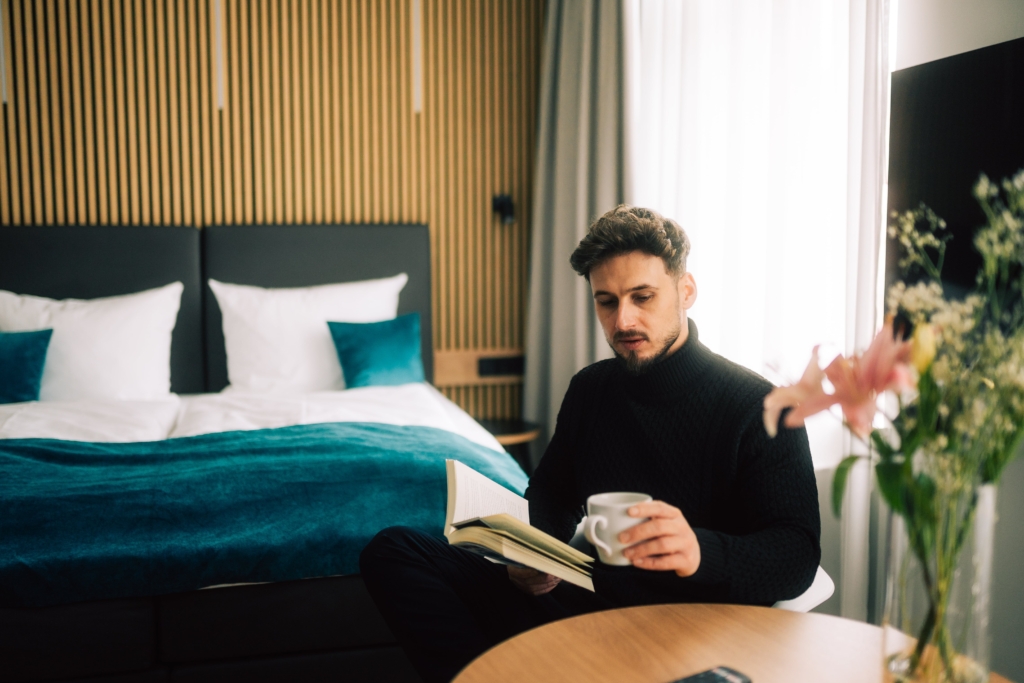 Ein Mann sitzt an einem Tisch in einem Zimmer im H24 Hotel Eberswalde, hält eine Tasse und ein aufgeschlagenes Buch in der Hand, im Hintergrund sind ein Bett und Blumen zu sehen.