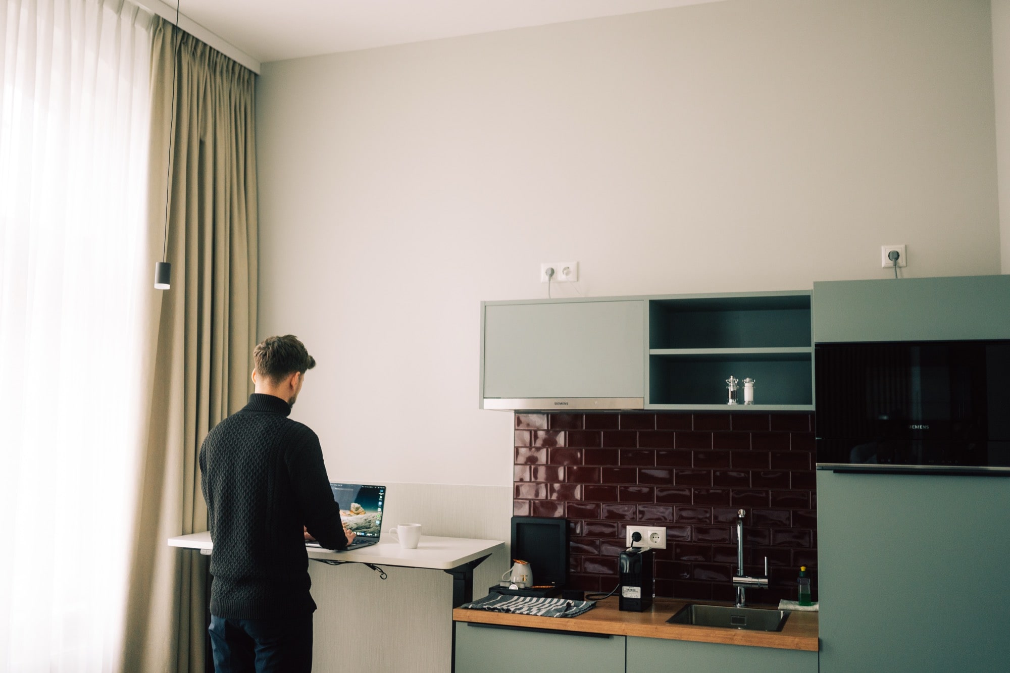 Eine Person steht am Tresen und arbeitet mit einem Laptop in einer modernen Küche mit hellen Schränken, roten Fliesen und natürlichem Licht durch ein Fenster mit langen Vorhängen.