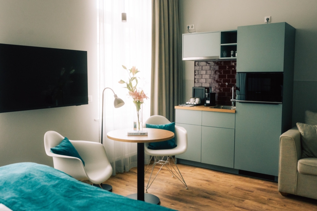 Modernes Studio-Apartment mit einer kleinen Küche, einem runden Tisch mit zwei Stühlen, einem an der Wand montierten Fernseher, einem Bett mit einer blauen Decke und einem Fenster mit durchsichtigen Vorhängen.