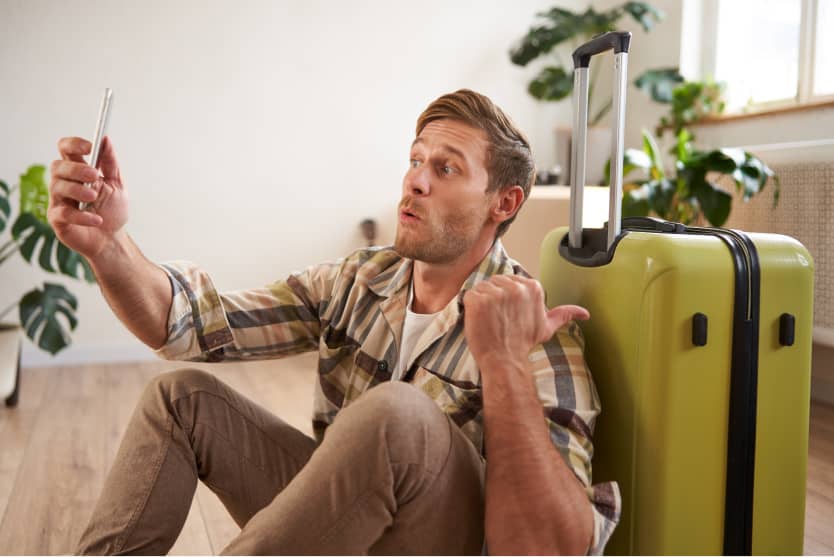 Mann macht Selfie, sitzt in Wohnung, grüner Koffer neben sich, Daumen nach oben