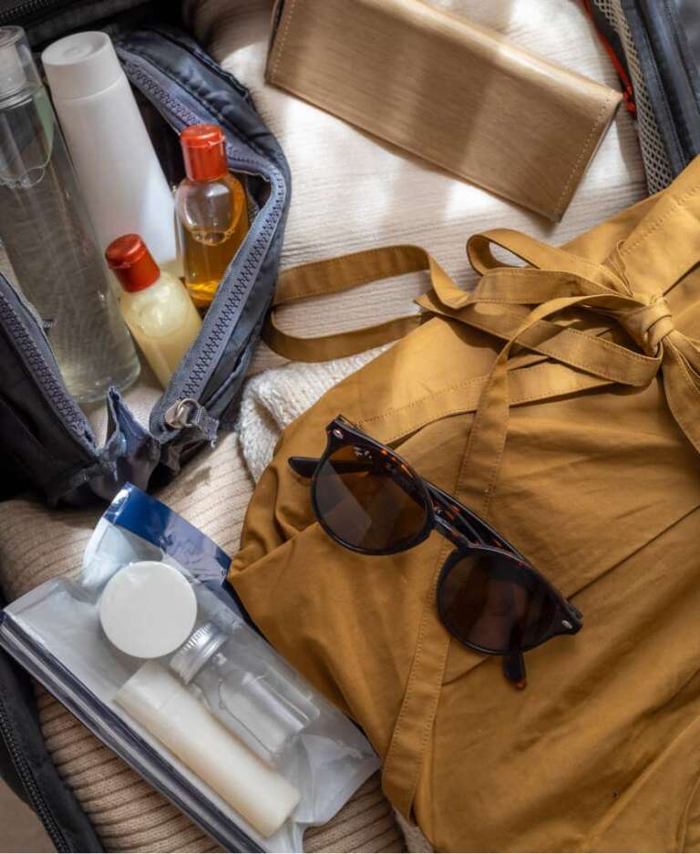 Geöffnete Reisetasche mit Kleid, Sonnenbrille und Kosmetiktasche mit Fläschchen innen