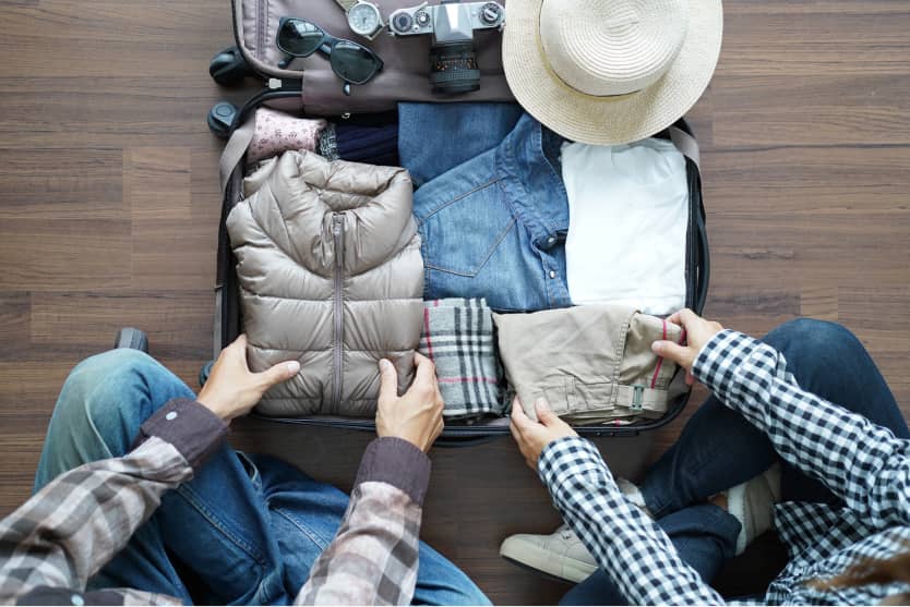 Zwei Personen packen gemeinsam Kleidung, Accessoires und Kamera in einen großen Koffer