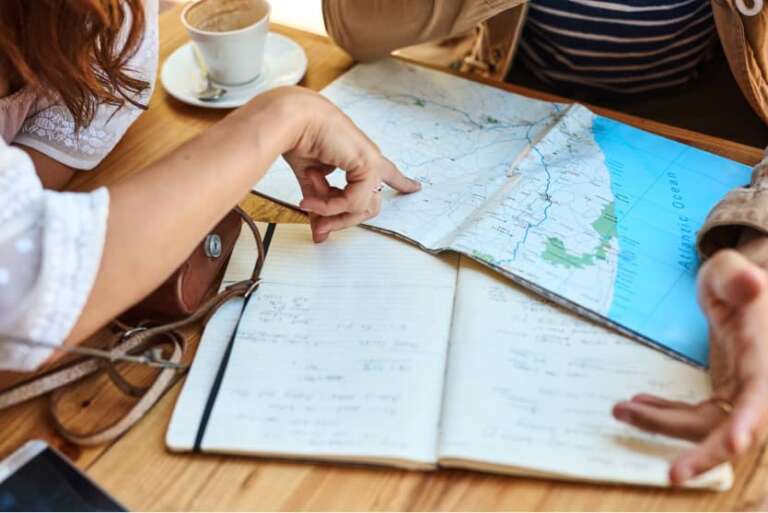 Zwei Personen sitzen am Tisch mit Karte, Notizbuch und Kaffee und planen gemeinsam Reise