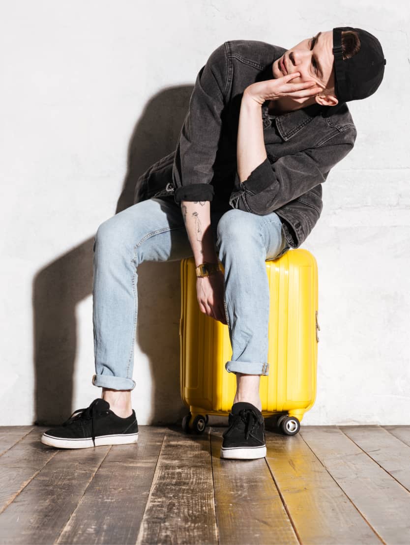 Junge Person sitzt nachdenklich auf gelbem Rollkoffer, trägt Jeans und dunkle Jacke