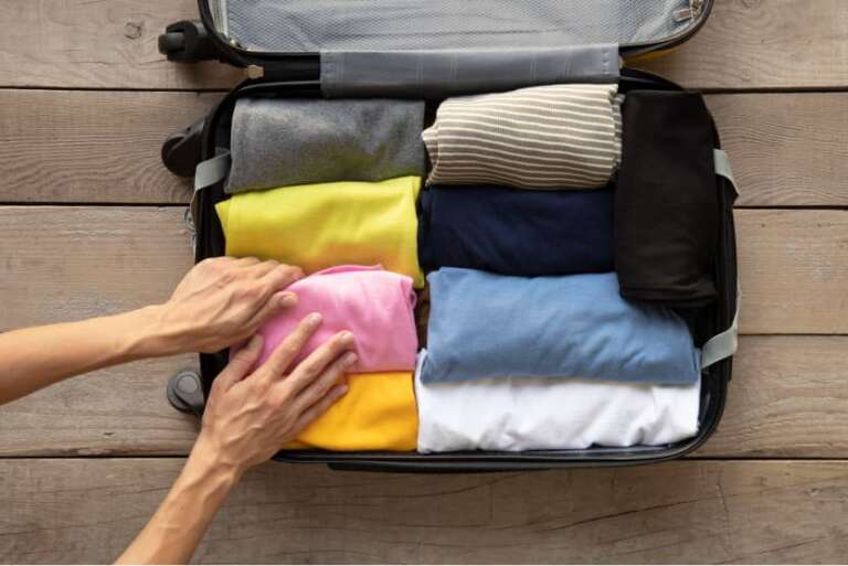 Offener Koffer auf Holzfußboden, ordentlich mit zusammengerollten Kleidungsstücken in verschiedenen Farben befüllt, eine Hand ordnet ein T-Shirt