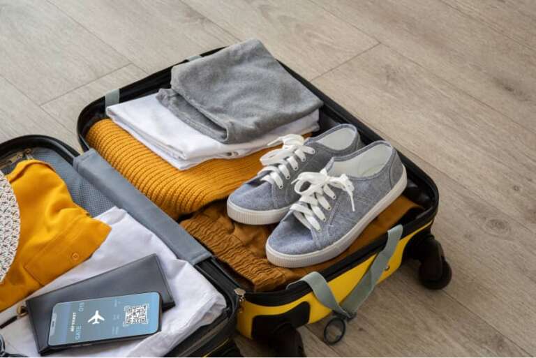 Gelber Reisekoffer auf Fußboden, darin gefaltete Kleidung, graue Turnschuhe und ein Smartphone mit digitaler Bordkarte.