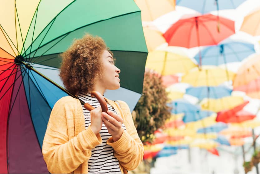 Person mit Regenbogen-Regenschirm bei bunten hängenden Schirmen, wetterfest gekleidet