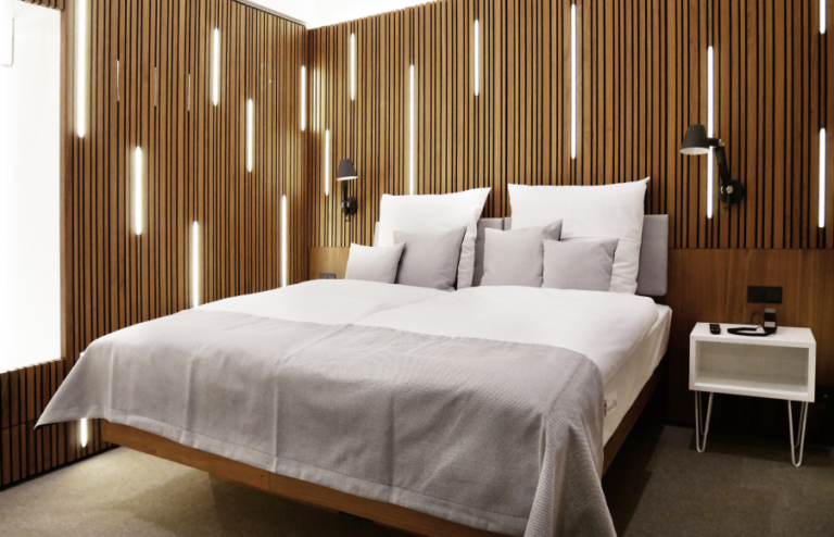 Ansicht eines modernen Zimmers mit Blick auf das Bett, einen Nachtschrank und Lichtpaneelen im Hintergrund. Ein Zimmer aus dem H24hotel in Bernau
