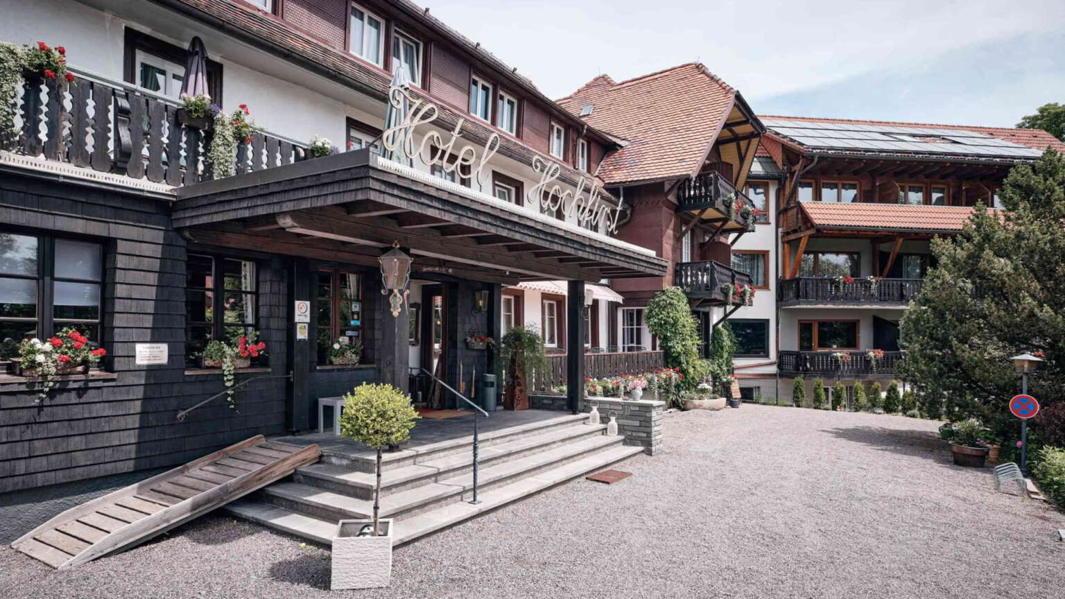 Hoteleingang vom H24 Hotel Hochfirst mit Treppe und Rampe, Topfpflanzen im Freien, Balkone mit Blumenkästen und Nebengebäude mit Holzelementen.