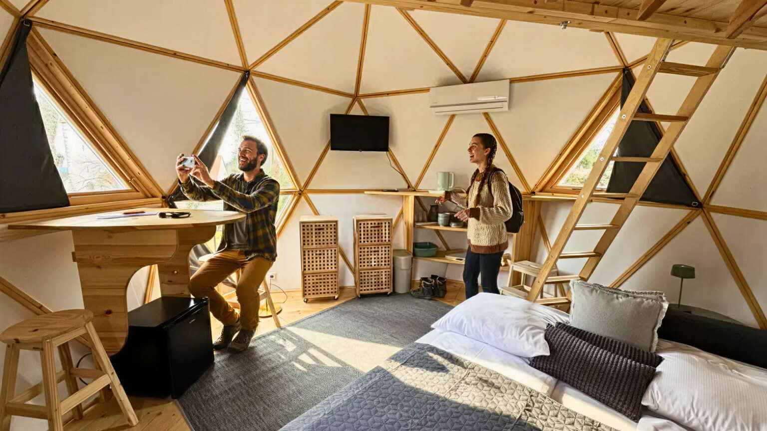 Zwei Personen stehen und sitzen in einer modernen geodätischen Kuppelkabine eines Tiny House, die mit Holzmöbeln, dreieckigen Fenstern und einem über eine Leiter erreichbaren Loft ausgestattet ist - ein innovativer Rückzugsort im H24 Hotel Hochfirst.
