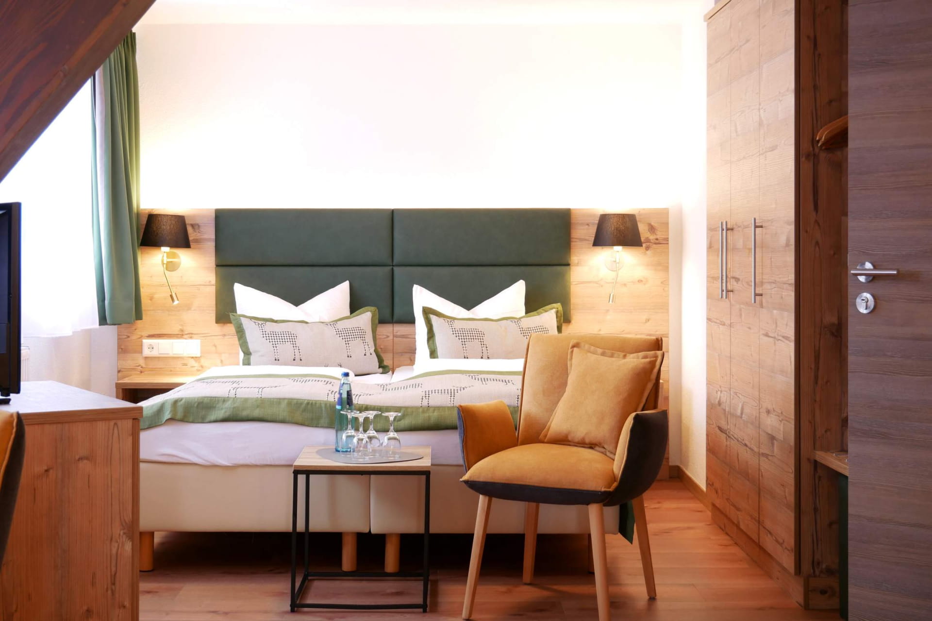 Ein ordentlich gemachtes Doppelbett mit grüner und weißer Bettwäsche, zwei Nachttischlampen, ein Stuhl mit Kissen, ein kleiner Tisch mit Wasser und Gläsern und ein hölzerner Kleiderschrank bilden ein gemütliches Schlafzimmer im Hotel Hochfirst.
