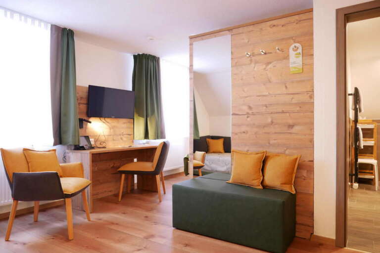 Modernes Hotelzimmer im Hotel Hochfirst mit Holzboden, Schreibtisch und TV, Sitzecke mit Stühlen und Sofa, grünen Vorhängen, Kleiderhaken und Blick in ein anderes Zimmer.