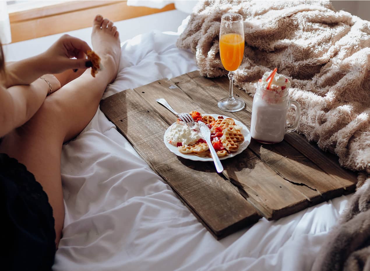 Junge Frau isst leckere Waffeln und trinkt köstliche Getränke auf einem Tablett auf dem Bett