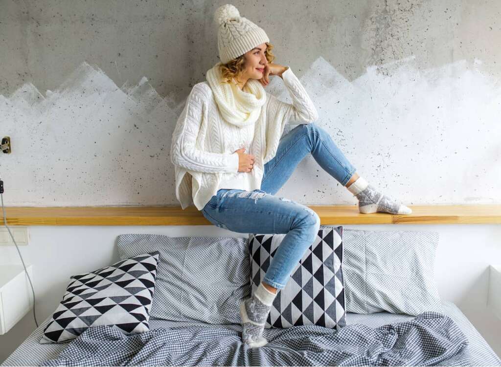 Person in Winterkleidung sitzt entspannt auf einem Bett mit geometrischen Kissen.
