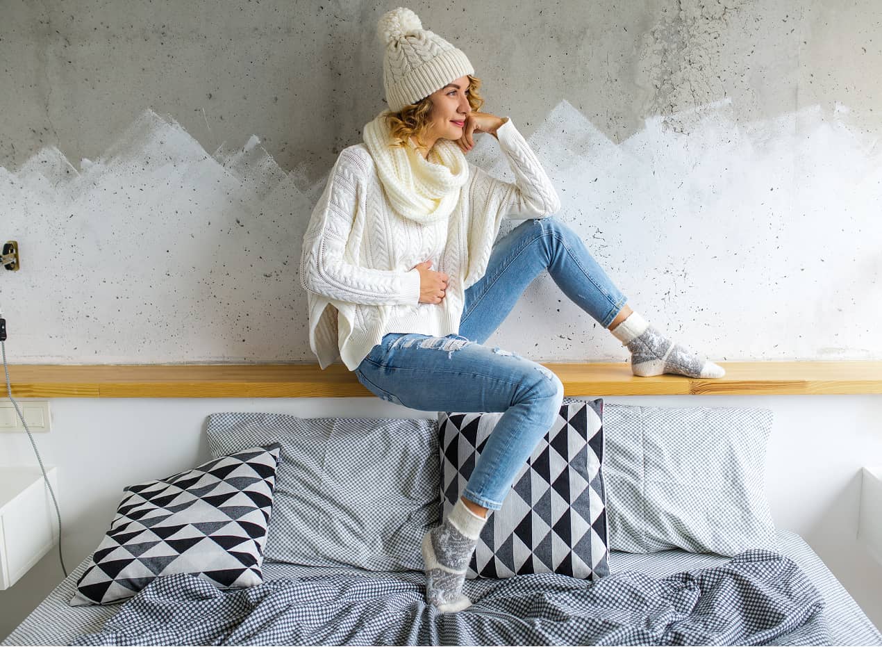 Person in Winterkleidung sitzt entspannt auf einem Bett mit geometrischen Kissen.