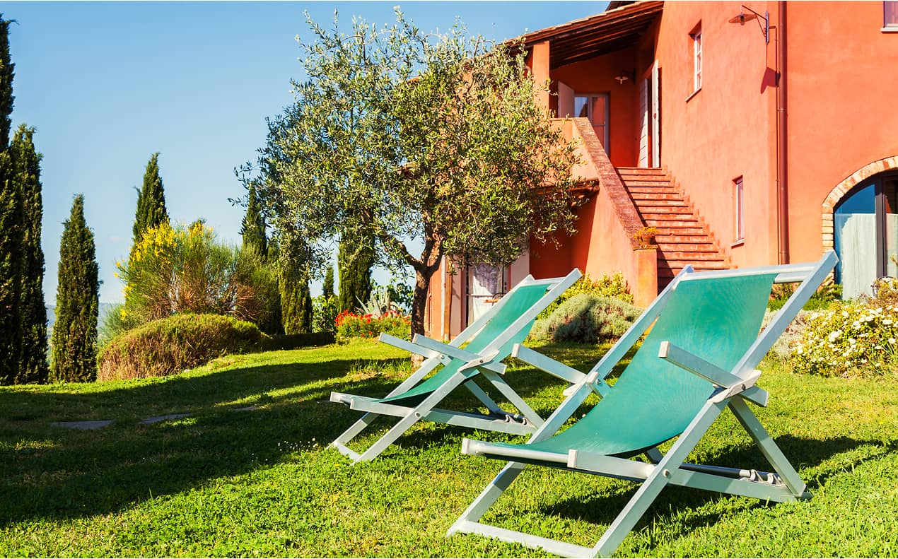 Zwei Liegestühle im Garten vor einem mediterranen Haus mit Bäumen und Blumen.
