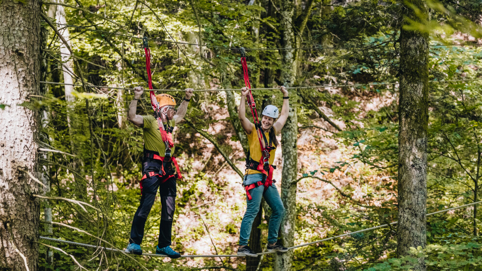 Mann und Frau mit Helmen und Klettergurten balancieren lachend über ein Drahtseil im Hochseilgarten, umgeben von grünem Laubwald.