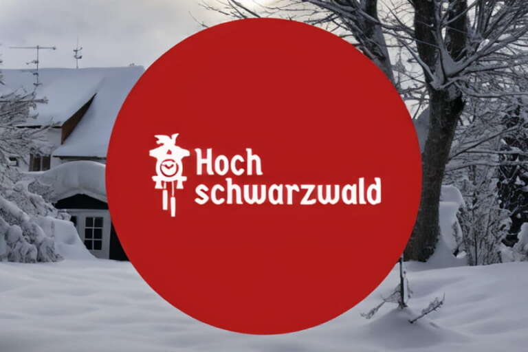 Ein roter Kreis mit dem weißen Hoch Schwarzwald-Logo überlagert eine verschneite Landschaft mit Bäumen und einem Haus im Hintergrund.