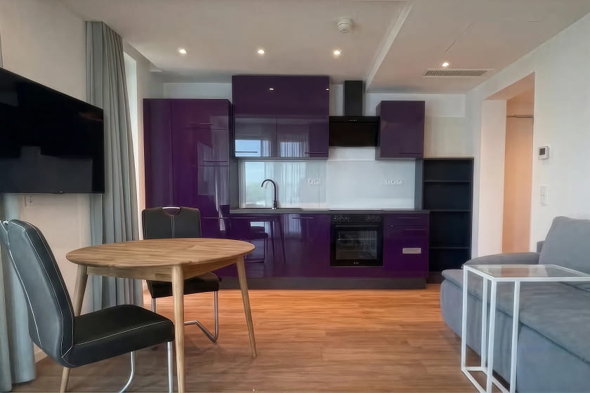 Moderne Küche und Wohnbereich mit glänzenden violetten Schränken, einem runden Holztisch mit zwei Stühlen, einem grauen Sofa, einem an der Wand montierten Fernseher und einem Holzboden.