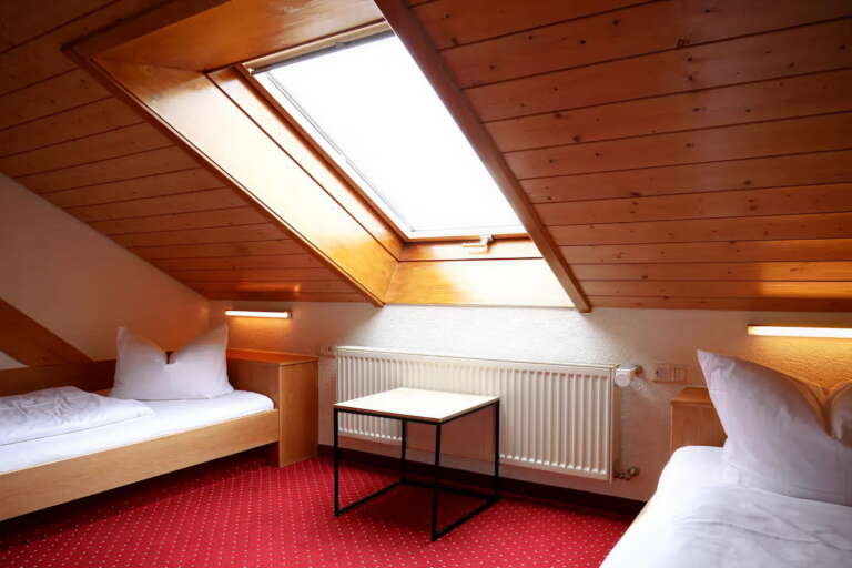 Zimmer im Dachgeschoss des Hotels Hochfirst mit zwei Einzelbetten, einem kleinen Tisch, rotem Teppich, holzgetäfelter Decke und einem Dachflächenfenster.