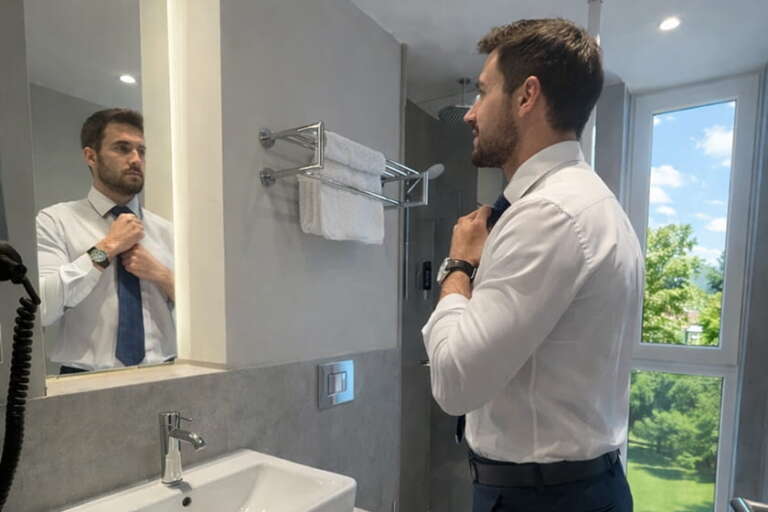 Ein Mann in einem weißen Hemd richtet seine Krawatte, während er sich im Badezimmerspiegel betrachtet.