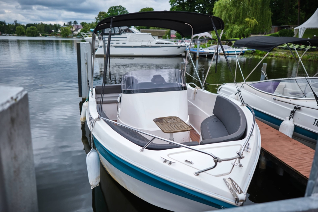 Ein weiß-blaues Motorboot des Bootsverleihs Berlin liegt an einer Anlegestelle auf einem ruhigen See. Im Hintergrund sind unter einem bewölkten Himmel in der Nähe des H24 DämeritzSeehotels weitere Boote zu sehen.