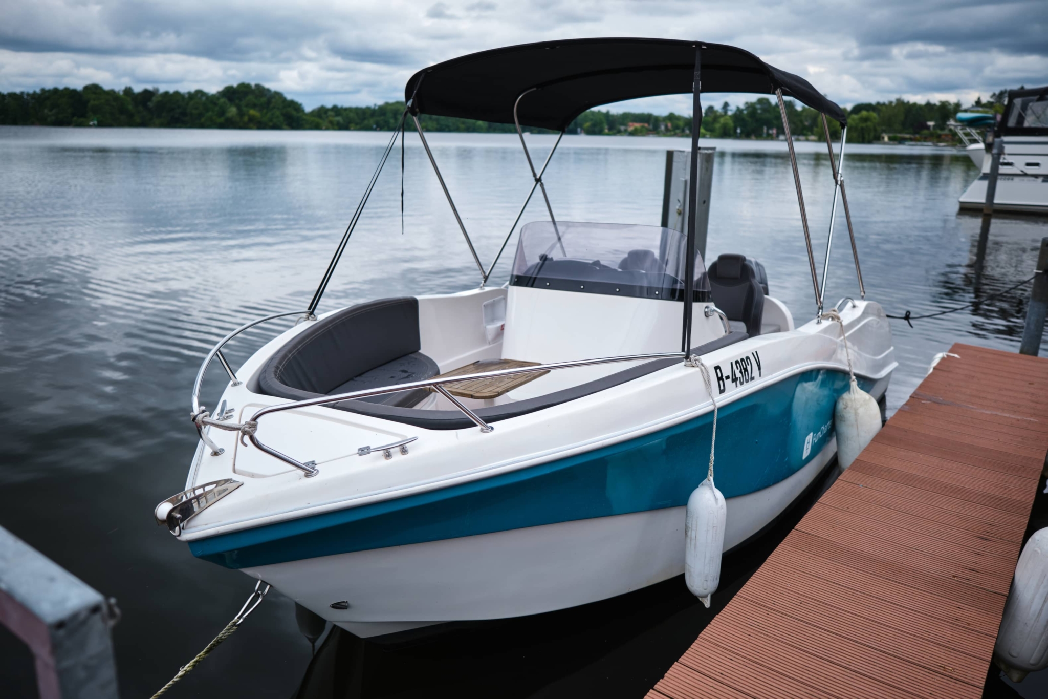 Ein kleines Motorboot mit blau-weißem Rumpf liegt an einer Anlegestelle auf einem ruhigen See in der Nähe des H24 DämeritzSeehotels. Es hat ein schwarzes Verdeck, Schutzfender an den Seiten und kann über den Bootsverleih Berlin gemietet werden.