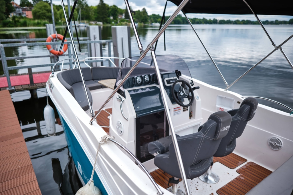 Ein kleines weißes Motorboot, das an einem Holzsteg am H24 DämeritzSeehotel angedockt ist. Es ist mit zwei schwarzen Kapitänssitzen, einem Steuerrad und Armaturenbrett-Bedienelementen ausgestattet, im Hintergrund sind Bäume und Wasser zu sehen. Bootsverleih Berlin bietet die perfekte Vermietung für einen malerischen Tag auf dem Wasser.