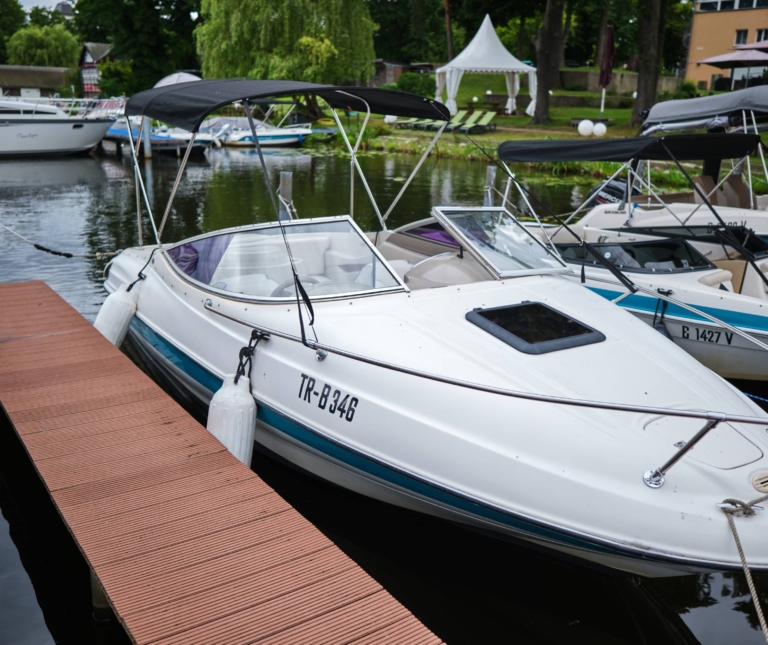 Ein weißes Motorboot mit der Kennzeichnung „TR-B346“ liegt an einem Holzsteg einer Marina, umgeben von mehreren anderen Booten auf einem ruhigen Gewässer, unweit des H24 DämeritzSeehotels.