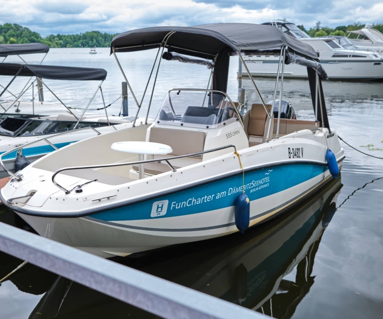 Ein Motorboot mit dem Logo „FunCharter am DämeritzSeehotel“, das neben anderen Booten in einem Yachthafen angedockt ist, bietet ein Vordach und einen Sitzbereich am Heck. Perfekt für Gäste des H24 DämeritzSeehotels oder für Nutzer des Bootsverleihs Berlin.