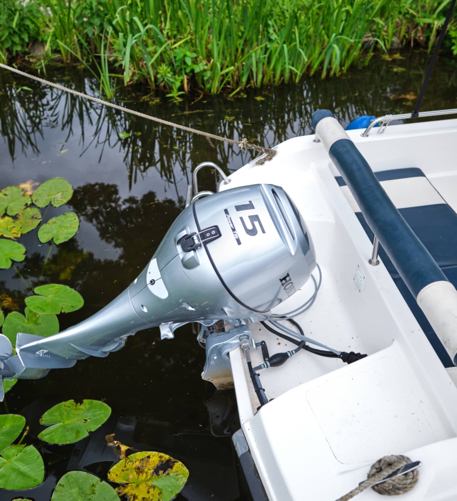 Ein 15-PS-Außenbordmotor ist am Heck eines kleinen weißen Bootes angebracht, das in einem mit Seerosen gefüllten Gewässer in der Nähe grüner Vegetation vertäut ist. Das Boot kann beim Bootsverleih Berlin beim malerischen H24 DämeritzSeehotel gemietet werden.