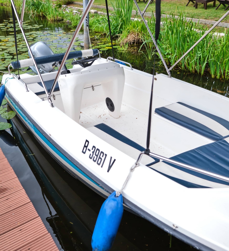 Ein kleines weißes Boot mit blauem Rand und dem Kennzeichen B-3961 V liegt an einem Holzsteg, umgeben von üppigem Grün, beim Bootsverleih Berlin, in der Nähe des H24 DämeritzSeehotels.