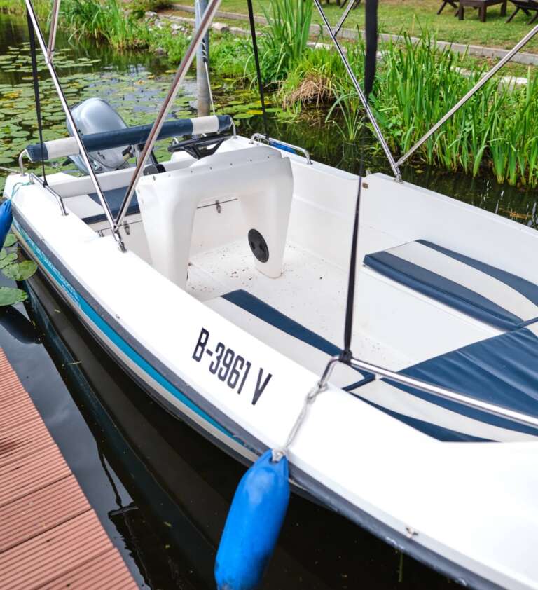 Ein kleines weißes Boot mit blauem Rand und dem Kennzeichen B-3961 V liegt an einem Holzsteg, umgeben von üppigem Grün.