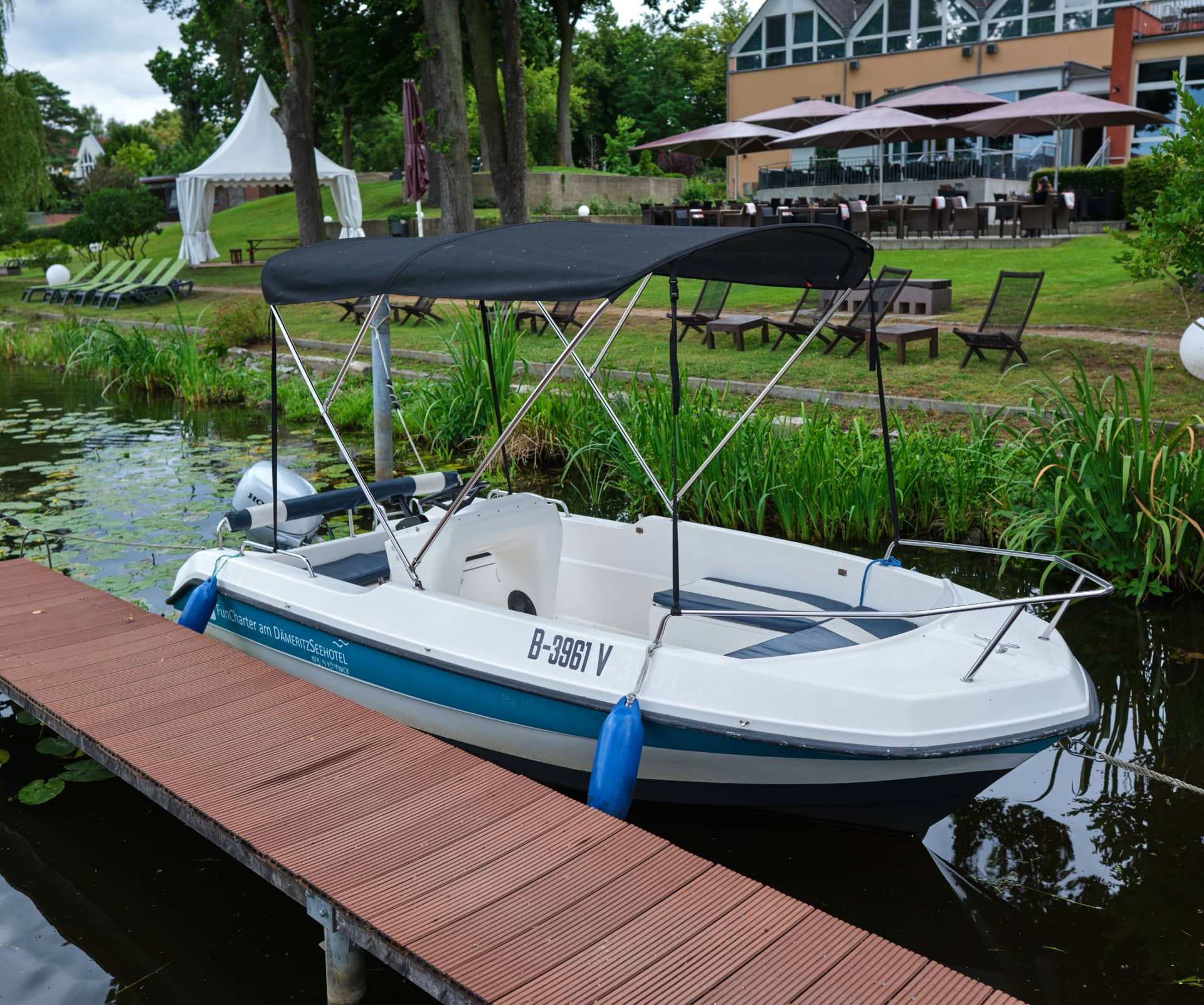 Mehrere weiß-blaue Boote liegen in einem Yachthafen in der Nähe des H24 DämeritzSeehotels. Eine Person steht auf dem Steg neben den Booten. Im Hintergrund sind Bäume und Gebäude zu sehen, die einen friedlichen Moment beim Bootsverleih Berlin einfangen.