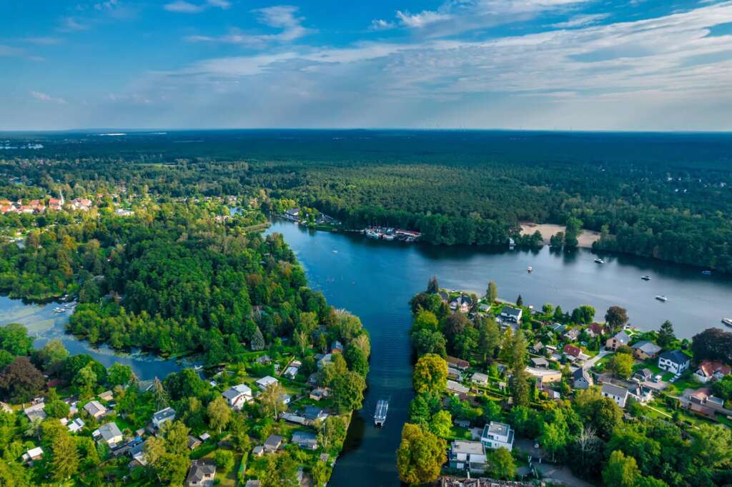 Luftaufnahme eines Flusses, umgeben von dichten grünen Wäldern und Wohngebieten, mit Häusern, Booten auf dem Wasser und einem wolkig-blauen Himmel in der Nähe von Bootsverleih Berlin und H24 DämeritzSeehotel.