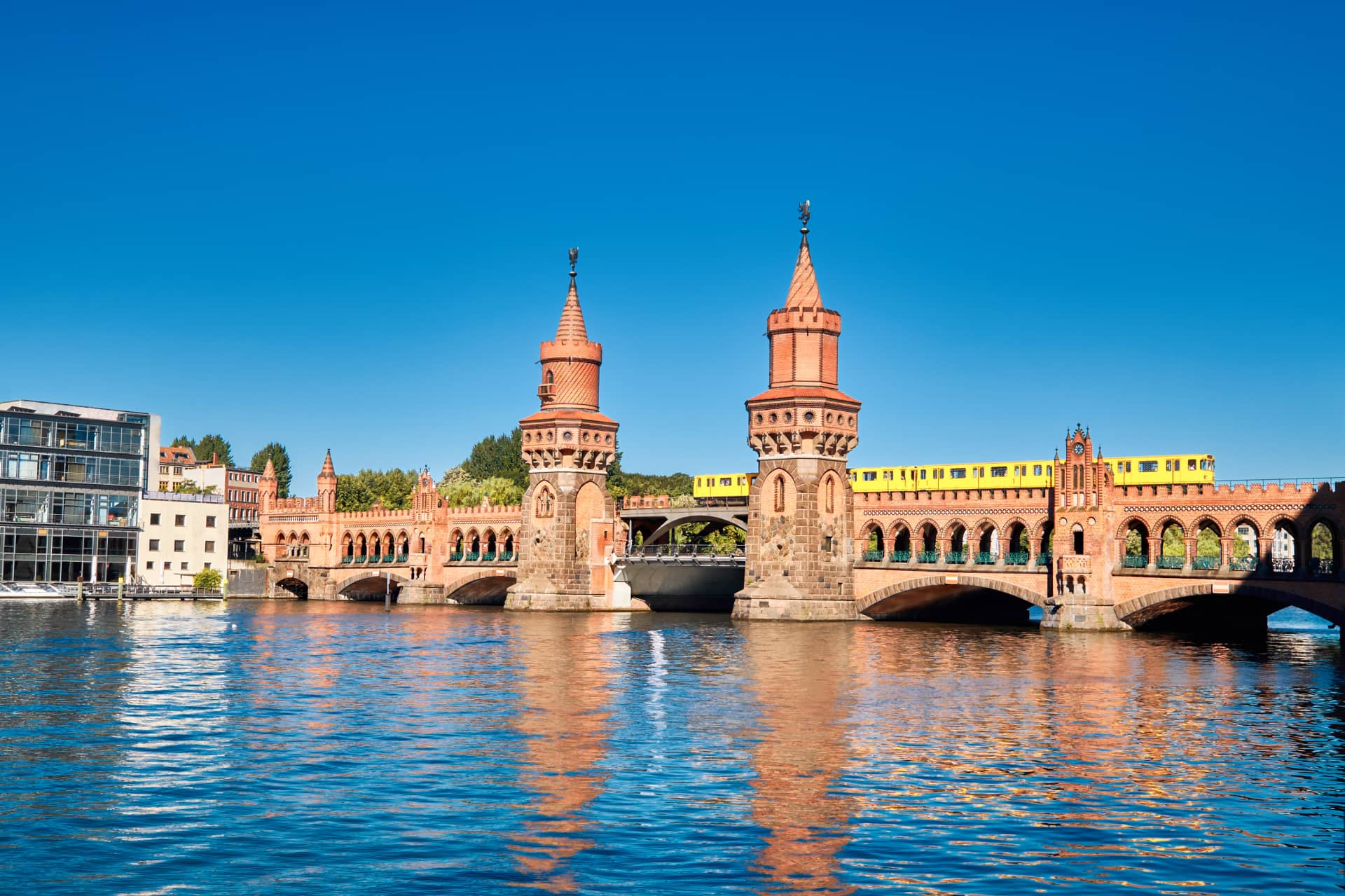 Eine rote Backsteinbrücke mit zwei Türmen überspannt einen Fluss unter einem klaren blauen Himmel. Ein gelber Zug fährt über die Brücke, links sind moderne Gebäude zu sehen. In der Nähe finden Sie den das H24 DämeritzSeehotel für einen längeren Aufenthalt oder einen Bootsverleih.