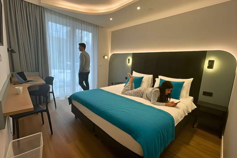 Eine Person steht am Fenster, während eine andere auf einem Bett liegt und ihr Handy benutzt – in einem modernen Hotelzimmer mit blauen Akzenten.