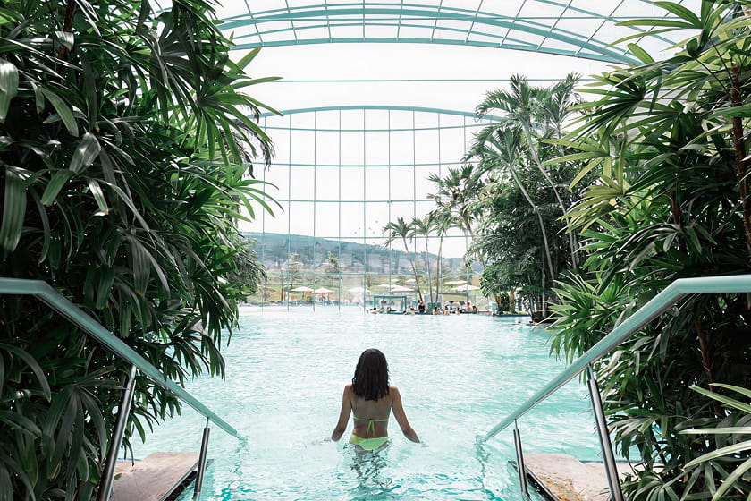 Eine Frau im Bikini betritt ein Hallenbad, das von tropischen Pflanzen umgeben ist, mit großen Glasfenstern und einem Blick ins Freie im Hintergrund.