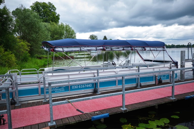 Ein überdachtes Pontonboot liegt an einem Holzsteg mit einem roten Teppichboden. Im Hintergrund sind grüne Bäume und Sträucher zu sehen, darüber ein bewölkter Himmel. Auf der Seite des Bootes, in der Nähe des H24 DämeritzSeehotels, ist eine Telefonnummer für Bootsverleih Berlin zu sehen.