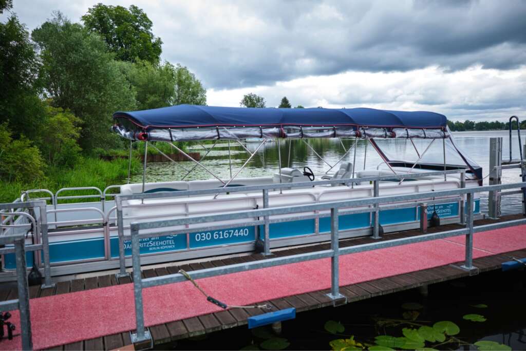 Ein überdachtes Pontonboot liegt an einem Holzsteg mit einem roten Teppichboden. Im Hintergrund sind grüne Bäume und Sträucher zu sehen, darüber ein bewölkter Himmel. Auf der Seite des Bootes, in der Nähe des H24 DämeritzSeehotels, ist eine Telefonnummer für Bootsverleih Berlin zu sehen.