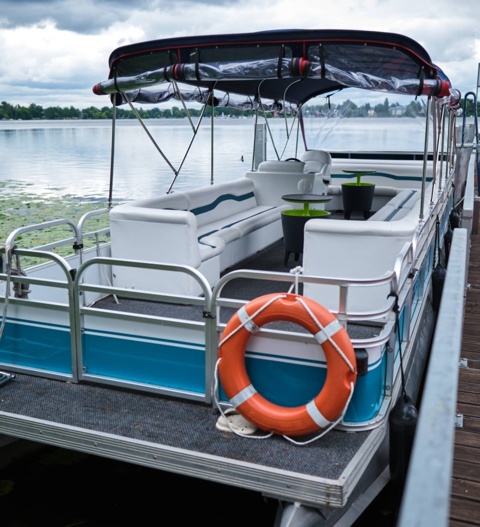 Auf einem ruhigen See unter wolkenverhangenem Himmel schwimmt ein angedocktes Pontonboot mit weißen Sitzen, einem Rettungsring an der Seite und einem Verdeck darüber, so wie man es beim Bootsverleih Berlin in der Nähe des H24 DämeritzSeehotels mieten kann.