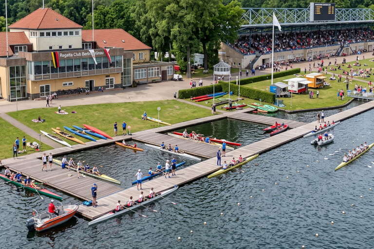Die Rudermannschaften bereiten sich an den Anlegestellen eines Regattavereins vor, während sich Zuschauer auf der Wiese und auf den Tribünen befinden und sich mehrere Menschen am Wasser versammelt haben.
