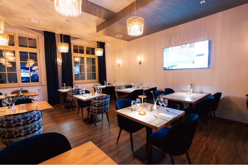 Modernes Restaurant im H24 Parkhotel in Thale mit Holztischen, blauen Stühlen, gemusterten Sesseln, Wandfernseher, großen Fenstern und hängenden Kronleuchtern, alles bei warmer Raumbeleuchtung.