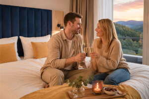 Ein Mann und eine Frau sitzen auf einem Bett in einem modernen Hotelzimmer, lächeln einander an und halten Gläser mit Champagner in den Händen; vor ihnen stehen ein Tablett, eine Kerze und Snacks, und draußen bietet sich ein malerischer Ausblick.