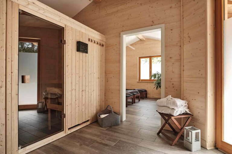 Moderne Sauna mit Holzwänden, einer Glastür, einer Bank mit gefalteten Handtüchern, einem Korb und einer Laterne; durch die offene Tür ist der angrenzende Raum zu sehen.